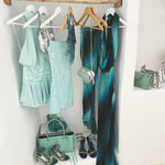 A styled closet display featuring a light blue lace dress, a teal gradient maxi dress, a mint green dress, and metallic silver accessories, including a handbag and shoes, hanging on a wooden rod and shelf.